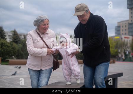 Kleines Mädchen, das mit Mann und Frau im Freien läuft. Großvater und Großmutter helfen der Enkelin beim Gehen Stockfoto