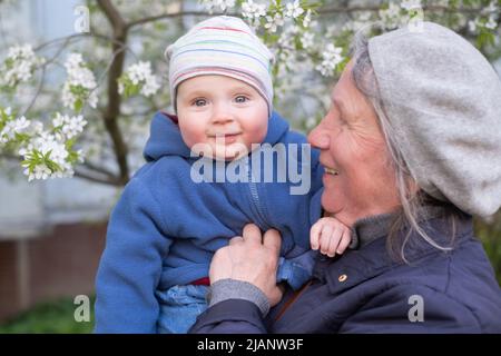 Ältere Großmutter, die ein 9 Monate altes Baby in den Armen hält und auf der Straße in der Nähe eines blühenden Baumes steht. Stockfoto