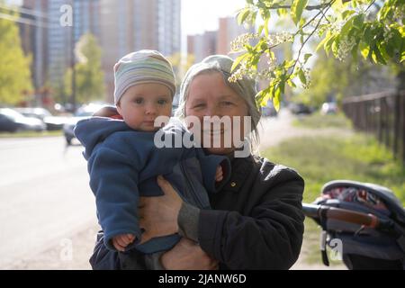 Ältere Großmutter, die ein 9 Monate altes Baby in den Armen hält und auf der Straße in der Nähe eines blühenden Baumes steht. Stockfoto