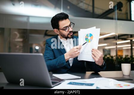Beschäftigter junger islamischer Geschäftsmann mit Bart in Brille und Anzug zeigt Diagramm zum Laptop im Büro Stockfoto