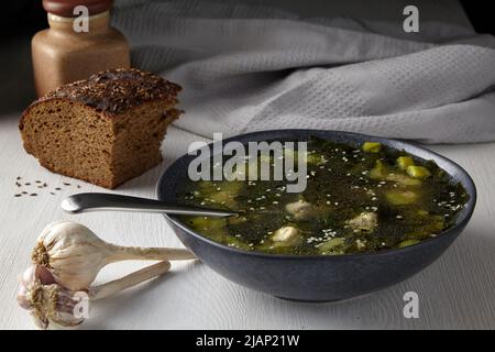 Rustikales Mittagessen im Licht vom Fenster. Zucchini-Suppe mit Fleischbällchen mit einem Stück schwarzem Brot mit Kümmel auf einem weißen Tisch gegen die BA Stockfoto