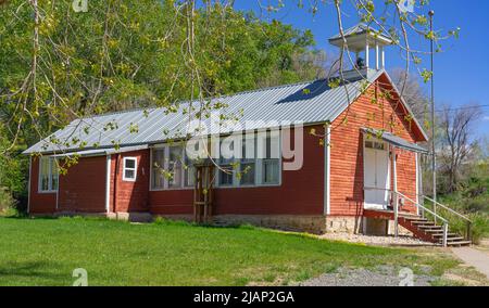 Das alte rote Schulhaus in Shell, WY, hat einen leeren Glockenturm und Holztreppen und Geländer, die zu den geschlossenen Eingangstüren führen. Es liegt auf einem grünen Hügel. Stockfoto