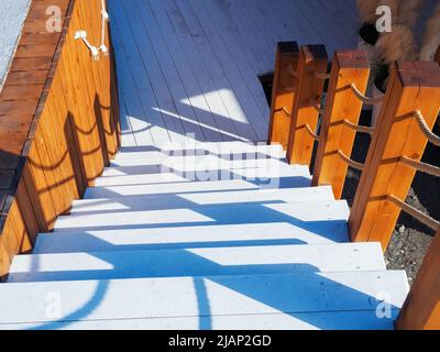 Weiße Holztreppen mit dekorativen, von der Sonne beleuchteten Geländern im nautischen Stil führen hinunter in einen schattigen Bereich. Stockfoto