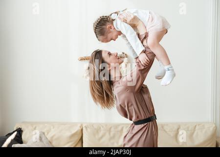 Lächelnde Mutter hebt die glückliche kleine Tochter im Wohnzimmer in die Luft und spielt mit ihr Stockfoto