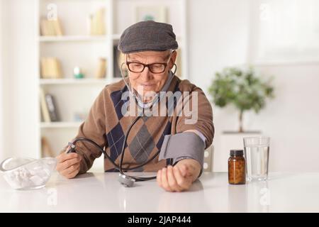 Älterer Mann, der den Blutdruck zu Hause mit Pillen in einer Flasche auf einem Tisch überprüft Stockfoto