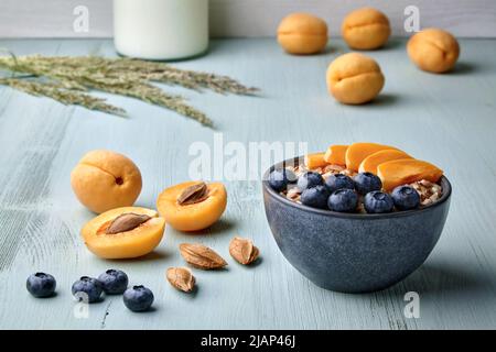 Vollkorn-Haferflocken mit Leinsamen, Aprikosenstücken und Heidelbeeren auf einem blauen Holztisch, umgeben von Zutaten. Gesundes Frühstück für Energie Stockfoto