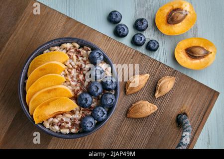 Vollkorn-Haferflocken mit Aprikosenstücken und Heidelbeeren auf einem Holztablett auf einem bemalten blauen Holztisch. Veganes Frühstück. Draufsicht. Flach liegend Stockfoto