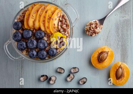 Vollkorn-Haferflocken mit Aprikosenstücken und Heidelbeeren und Leinsamen werden auf einem bemalten blauen Holztisch mit Blumen garniert. Welt-Vegetarier-Tag Stockfoto