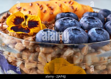 Vollkorn-Haferflocken mit Aprikosenstücken und Heidelbeeren und Leinsamen werden mit Blumen garniert. Welt-Vegetarier-Tag Stockfoto