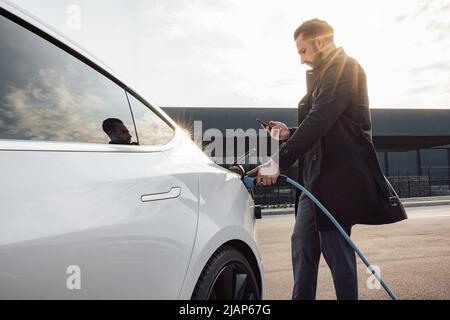 Ein eleganter Geschäftsmann, der bei Sonnenuntergang ein neues weißes Elektroauto auf dem Parkplatz des Handelszentrums lädt Stockfoto