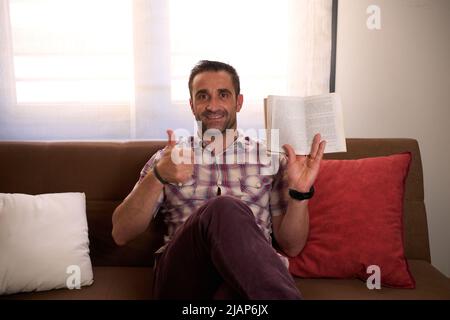 Mann in einem karierten Hemd und Buch in der Hand, entspannt auf einem Sofa sitzend Stockfoto