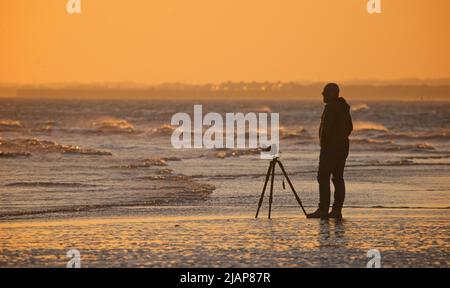 Fotograf mit Kamera auf Stativ, tragbar, bei Ebbe am Ufer, Brighton, Sussex, England, Großbritannien. Sonnenuntergang/Abenddämmerung. Stockfoto