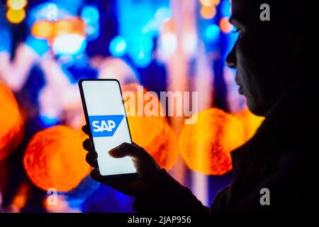 In dieser Abbildung hält eine silhouettierte Frau ein Smartphone mit dem SAP SE-Logo auf dem Bildschirm. Stockfoto