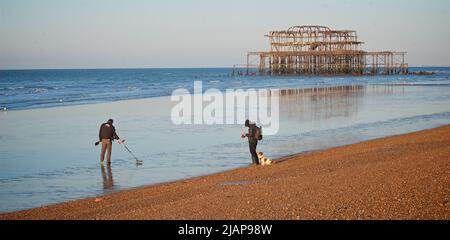 Rostreste des verfallenen West Pier bei Ebbe im Morgengrauen. Brighton & Hove, Sussex, England, Großbritannien. Metalldetektorin prospektiert den Sand und Mann mit Hund und Kamera am Kiesstrand. Stockfoto