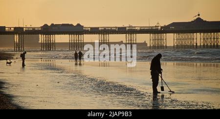 Metalldetektorin prospektiert zu wenig Zeit den Sand. Beach at Dawn, Brighton & Hove, Sussex, England, Großbritannien. Menschen, die auf dem Sand spazieren, einige mit einem Hund; Palace Pier dahinter. Stockfoto