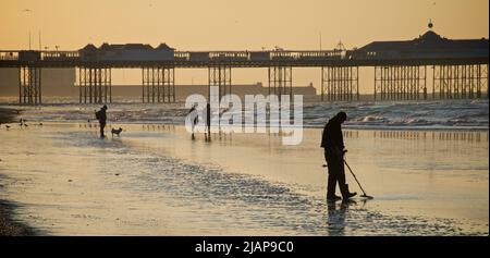 Metalldetektorin prospektiert zu wenig Zeit den Sand. Beach at Dawn, Brighton & Hove, Sussex, England, Großbritannien. Menschen, die auf dem Sand spazieren, einige mit einem Hund; Palace Pier dahinter. Stockfoto