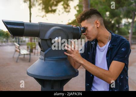 Ein junger Mann in legerer Kleidung, der neugierig durch ein Teleskop auf einem öffentlichen Platz aussieht. Stockfoto