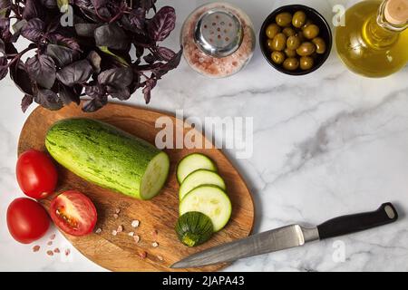 Zucchini-Tomaten Oliven Basilikumöl und Salz auf einem Holzbrett auf einem hellen Tisch. Zutaten für die Zubereitung vegetarischer Mahlzeiten Stockfoto