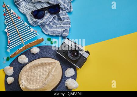 Strohhut, Sonnenbrille, Segelboot und Kamera auf gelb-blauem Hintergrund. Sommerurlaub Konzept auf See, Reisen Stockfoto