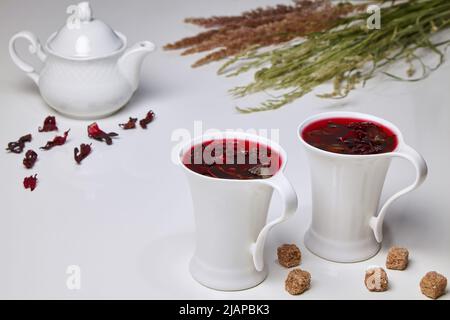 Hibiscus Kräutertee aus sudanesischen Rosen- und Minzblüten mit Rohrzucker-Zangen in eleganten weißen Tassen auf einem weißen Tisch vor dem Hintergrund Stockfoto