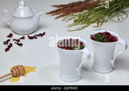 Hibiskus-Kräutertee aus sudanesischen Rosen- und Minzblüten in eleganten weißen Tassen auf einem weißen Tisch, umgeben von Honig, Teekanne und einem Bouquet aus Wild Stockfoto