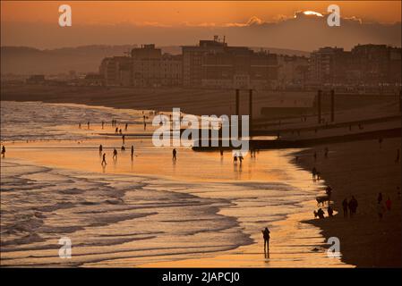 Silhouetten von Menschen am Strand bei Ebbe, Brighton & Hove, East Sussex, England, Großbritannien. Die Sonne geht hinter der Wolke auf der rechten Seite unter. Vertikale gusseiserne Säulen des alten West Pier auf der rechten Seite. Stockfoto