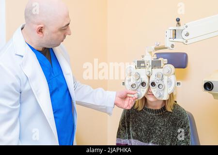 Ein männlicher Augenarzt überprüft das Augenlicht eines Mädchens mit einem Phoropter. Sehbehandlung. Stockfoto