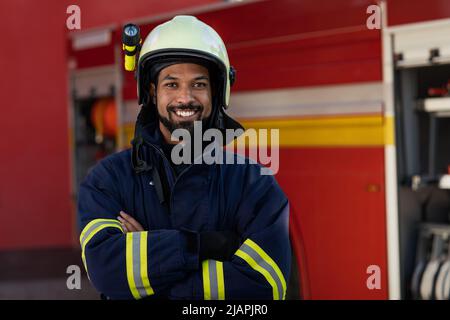 Glücklicher junger afroamerikanischer Feuerwehrmann mit Feuerwehrauto im Hintergrund. Stockfoto
