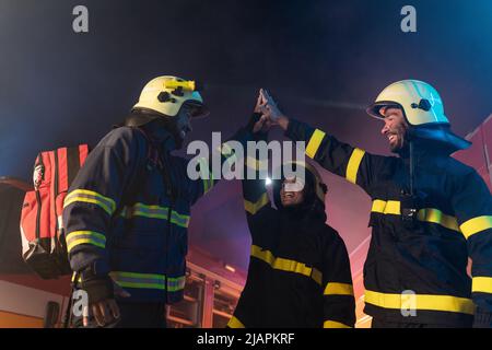 Glückliche Feuerwehrleute Männer und Frau nach erfolgreicher Aktion mit Feuerwehrauto im Hintergrund in der Nacht Stockfoto