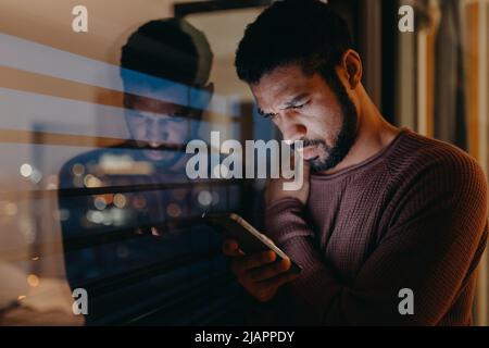 Ein junger Mann mit Smartphone sieht besorgt aus und steht abends zu Hause auf dem Balkon. Stockfoto