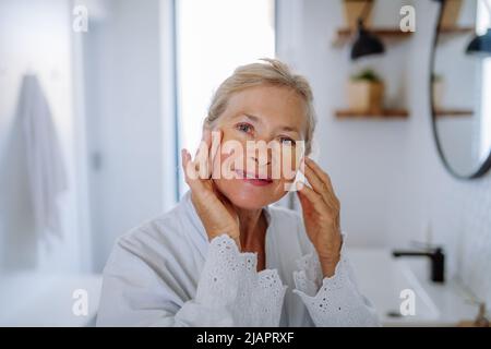 Schöne ältere Frau im Bademantel, die Augenflecken auf Schwellungen aufsetzt, während sie die Kamera anschaut Stockfoto