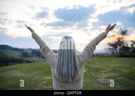 Rückansicht einer älteren Frau mit ausgestreckten Armen und dem Gesicht nach oben im Park am Frühlingstag Stockfoto