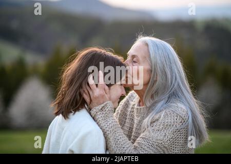 Glückliche ältere Großmutter, die ihren Teenager-Großdaguchter am Frühlingstag in der Natur auf die Stirn küsst. Stockfoto