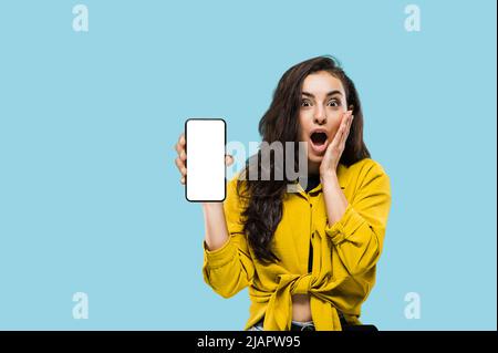 Betäubt schockiert kaukasische langhaarige Brünette Mädchen, in einem lässigen Hemd, schaut überrascht auf die Kamera, zeigt Smartphone mit leeren weißen Mock-up-Bildschirm, steht auf isoliertem blauen Hintergrund Stockfoto