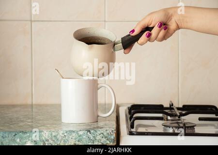 Eine Frauenhand gießt Kaffee aus einem türkenkaffee in eine Tasse auf dem Tisch in der Küche und bereitet den Morgenkaffee in der Küche zu Stockfoto