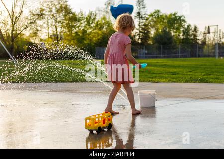 Kleines Mädchen, das im Sommer auf dem Spielplatz im Park mit Spielzeug spielt. Kleines Kind von hinten in der Nähe von Brunnen, das Zeit im Freien verbringt Stockfoto