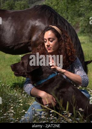 Weibchen mit ihrer Schokolade labrador Retriever in den Bergen in Frühlingswiese und schwarzes Pferd im Hintergrund. Stockfoto