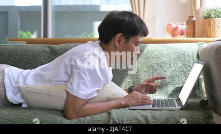 Junger asiatischer Mann in legerer Kleidung, der auf dem Sofa liegt und auf einem Laptop im Internet surft Stockfoto