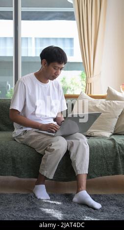 Ganzkörperaufnahme eines jungen Mannes, der im hellen Wohnzimmer mit einem Computer-Laptop sitzt Stockfoto