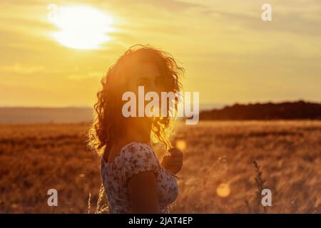 Junge marokkanerin mit braunen lockigen Haaren steht auf einem Weizenfeld, während die Sonne im Hintergrund untergeht und die Kamera blendet Stockfoto