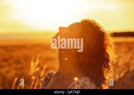 Junge marokkanerin mit braunen lockigen Haaren steht auf einem Weizenfeld, während die Sonne im Hintergrund untergeht und die Kamera blendet Stockfoto