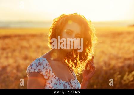 Junge marokkanerin mit braunen lockigen Haaren steht auf einem Weizenfeld, während die Sonne im Hintergrund untergeht und die Kamera blendet Stockfoto