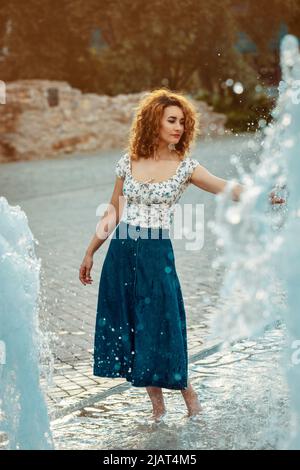 youn marokkanerin, mit lockigen braunen Haaren, trägt einen jean-Rock und spielt mit Wasser an einem Brunnen Stockfoto