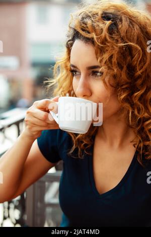 Junge marokkanerin mit lockigen braunen Haaren sitzt in einem Mainzer Café im Freien und trinkt Kaffee Stockfoto