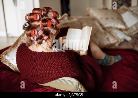 Rückansicht einer älteren Frau, die bunte Lockenwickler aufträgt, weinige Strickjacke trägt, auf dem Sofa sitzt und Buch liest. Stockfoto