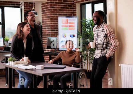 Eine vielfältige Gruppe von Menschen, die in der Pause am Arbeitsplatz Spaß haben, lachen und sich mit einem gelähmten Mitarbeiter unterhalten. Frau, die bei der Arbeit im Rollstuhl mit körperlicher chronischer Behinderung lebt. Stockfoto