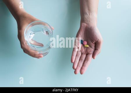 Hand hält Omega 3, Spirulina, Chlorophyll, Magnesiumkapseln über Glas Wasser auf Holztisch. Morgens Vitamintabletten. Nahrungsergänzungsmittel. Heilen Stockfoto