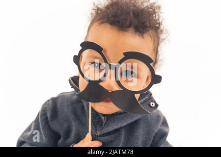 Mittlere Nahaufnahme über weißem Hintergrund eines interracial niedlichen Kleinkind Baby junge mit schwarzem gefälschten Papier Schnurrbart und Brille. Glückliche Kindheit Konzept. Hochwertige Fotos Stockfoto