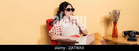 Stilvolle asiatische Frau in Sonnenbrille sitzt auf Sessel neben Telefon auf Couchtisch auf orangefarbenem Hintergrund, Banner Stockfoto