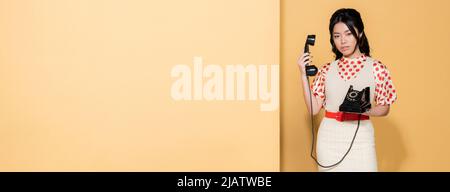Junge asiatische Modell in Retro-Kleidung mit Telefon auf orangefarbenem Hintergrund, Banner Stockfoto